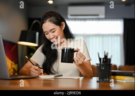 Cheerful young Asian female college student or freelancer working in the living room, sipping morning coffee while writing something on her notebook. Stock Photo