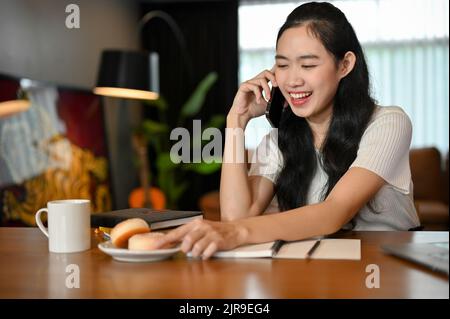 Attractive and cheerful young Asian female college student enjoys talking on the phone with someone while doing homework in her living room. Stock Photo