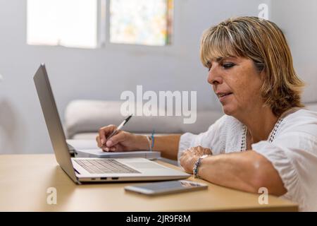 Mature woman sitting and working from home, jotting down notes in her diary and using her laptop with her cell phone next to it. Stock Photo