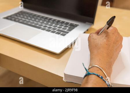 unrecognizable mature woman writing in her diary with her laptop next to her. Stock Photo