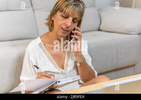 Adult woman working from home talking on the phone while taking notes in her diary. Stock Photo