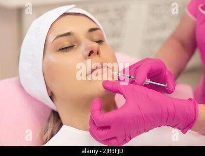 Top view of young woman lying on couch with closed eyes getting rejuvenation lifting injection into lips. Cosmetology. Stock Photo