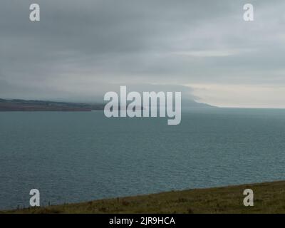 A heavy fog bank rolls over headland on the horizon, behind a intricate expanse of sea, under a huge, grey sky. Stock Photo