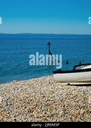 A small boat pulled onto a deserted shingle beach near a marker buoy and a rippled blue sea, under a clear summer sky. Stock Photo