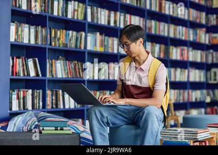 Portrait of young Asian man using laptop in library lounge with vibrant blue tones, copy space Stock Photo