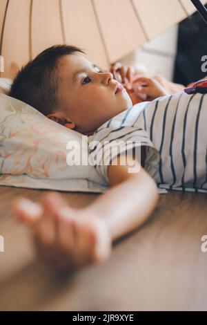 Adorable boy lying on his back on the playroom floor under the umbrella with his sister to put her to sleep. People lifestyle concept. Childhood Stock Photo