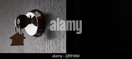 Key with a trinket in the form of a house in the door lock of an open door against the dark background Stock Photo