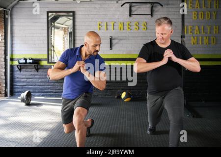 Male personal trainer instructing and assisting mature senior man while doing forward leg lunges dynamic movement exercise in home gym small business Stock Photo