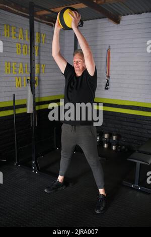 Mature man exercising in home garage gym. Senior man doing weighted medicine ball smashes exercise. Stock Photo