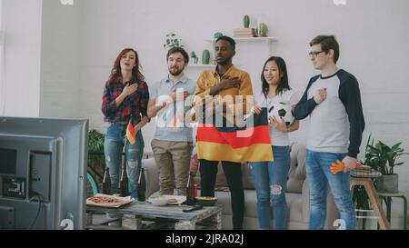 Multi-ethnic group of friends sport fans listening and singing German national anthem before watching sports championship on TV together at home indoors Stock Photo