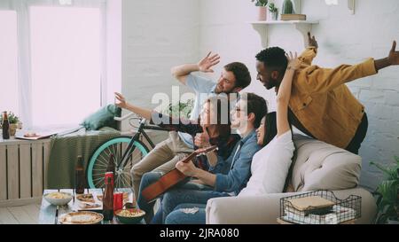 Multi ethnic group of cheerful friends taking selfie photos on smartphone camera while celebrating at party with beer and snacks at home Stock Photo