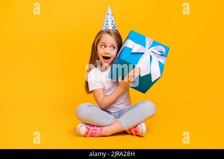 Full length photo of impressed suspicious school girl wear white t-shirt birthday cap listening inside present isolated yellow color background Stock Photo