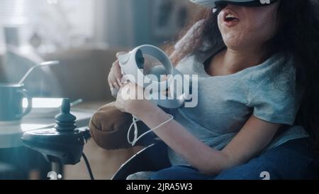 Woman with disability in a wheelchair at home wearing virtual reality glasses and holding controllers in her hands join metaverse Stock Photo