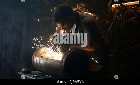 A male worker processing metal details for a bbq grill in a workshop for his small business wearing protective uniform, goggles and gloves Stock Photo