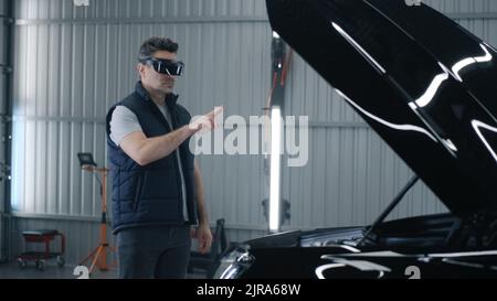 A male mechanic in a virtual reality headset gesturing and standing near the open hood of a car, while making a diagnostics in a car service Stock Photo