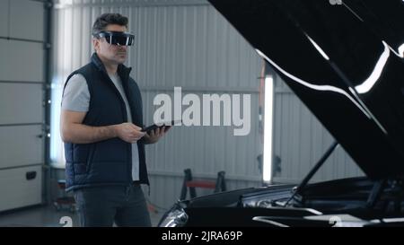 A male worker diagnosing and checking a car after a repairing in a car service using a tablet and a modern virtual reality glasses Stock Photo