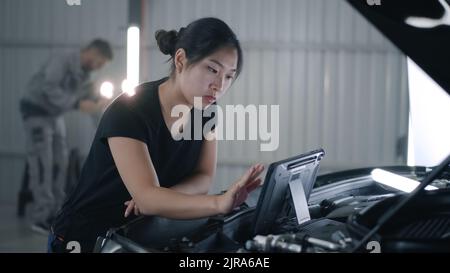 Asian female mechanic in uniform checking car engine and swiping on tablet computer during diagnostics and maintenance in car service Stock Photo