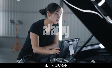 Asian female mechanic in uniform checking car engine and swiping on tablet computer during diagnostics and maintenance in car service Stock Photo