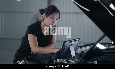 Asian female mechanic in uniform checking car engine and swiping on tablet computer during diagnostics and maintenance in car service Stock Photo