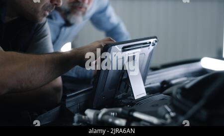 Car service mechanic clicking on a screen of digital tablet computer while standing next to a client, near the opened bonnet of the car during diagnostics and maintenance Stock Photo