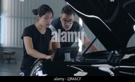 Male and female employees near the hood diagnosing and checking a car together in a car service using a tablet and discussing repairing Stock Photo