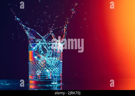 Whiskey glass with ice cubes and water splashes standing on mirror surface over gradient black orange background in neon light. Art, beauty, drinks Stock Photo