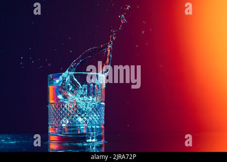 Whiskey glass with ice cubes and water splashes standing on mirror surface over gradient black orange background in neon light. Art, beauty, drinks Stock Photo