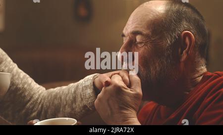 Elderly man kissing hand of happy woman and paying compliment while relaxing on couch and enjoying fresh tea in weekend at home Stock Photo