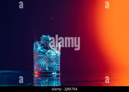 Ice cube falls into a transparent glass of water standing on mirror surface over gradient black orange background in neon light. Art, beauty, drinks Stock Photo