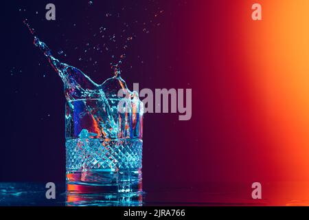 Closeup glass with ice cubes and water splashes standing on mirror surface over gradient black orange background in neon light. Art, beauty, drinks Stock Photo