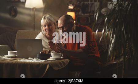 Aggressive man having argument with kids online, while sitting on sofa near woman and making video call via netbook laptop in cozy living room at home Stock Photo