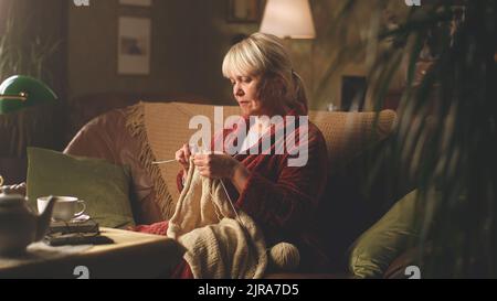 A retired woman knitting a warm sweater at her leisure time while sitting on the sofa in the evening near her husband with newspaper Stock Photo