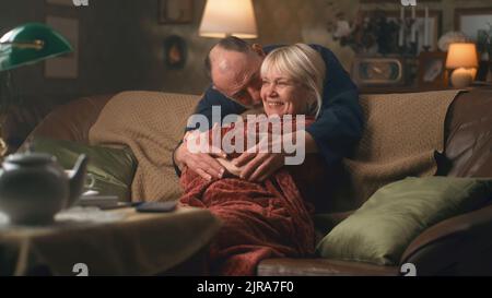 Aged husband embracing and kissing surprised wife in bathrobe resting on sofa in living room in evening at home Stock Photo