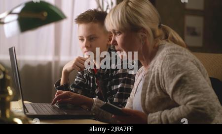 Teenager in checkered shirt showing grandmother with smartphone how to use netbook while sitting on sofa at home Stock Photo