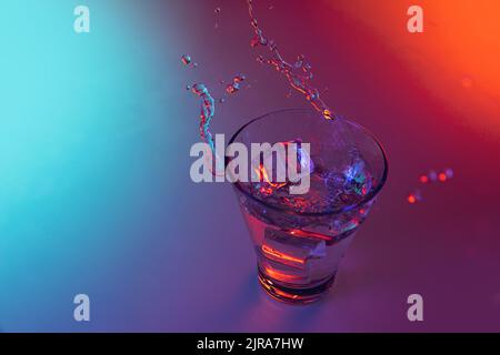 Closeup ice cube falls into a transparent glass of water standing on mirror surface over gradient red orange background in neon light. Art, beauty Stock Photo