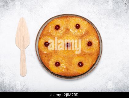 Homemade pineapple upside down cake with glace cherries and  wooden pie server on light rustic background. Top view. Copy space. Stock Photo