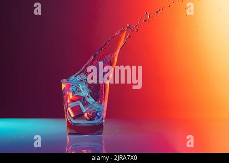 Ice cube falls into a transparent glass of water standing on mirror surface over gradient red orange background in neon light. Art, beauty, drinks Stock Photo