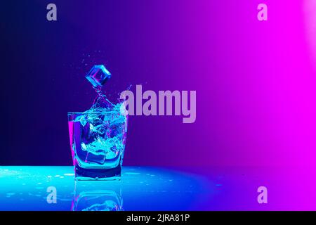 Ice cube falls into a transparent glass of water standing on mirror surface over gradient blue-pink background in neon light. Art, beauty, drinks Stock Photo