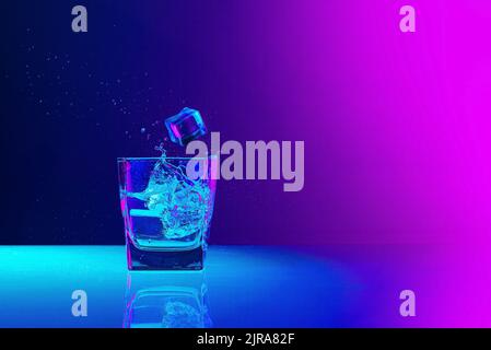 Ice cube falls into a transparent glass of water standing on mirror surface over gradient blue-pink background in neon light. Art, beauty, drinks Stock Photo
