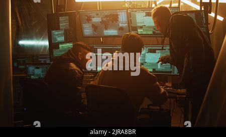 Young man in hoodie helping associate to hack database for criminal while gathering around table in dim room Stock Photo
