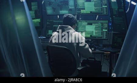 Happy young man in hoodie with glasses and hat dancing in excitement after successful hacker attack, while sitting at desk with computers in dark room Stock Photo