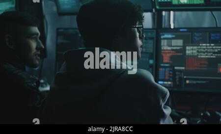 Young male hacker in glasses looking at screen and talking with associate while performing cyber attack on computer in dim room of criminal base Stock Photo