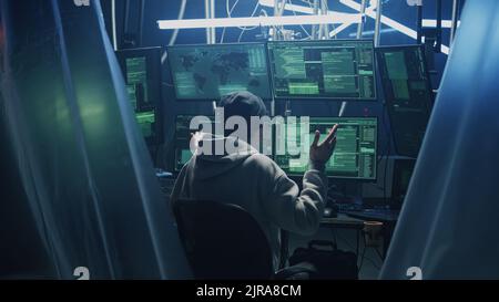 Happy young man in hoodie with glasses and hat dancing in excitement after successful hacker attack, while sitting at desk with computers in dark room Stock Photo