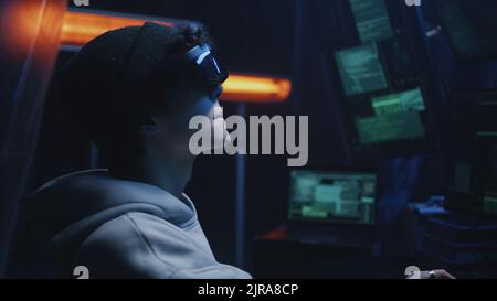 A teen hacker in a hat and augmented reality glasses siting at a desk in cybercriminal refuge near computers Stock Photo