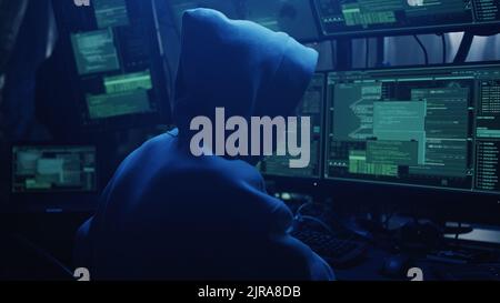 Anonymous hacker in hoodie writing code and explaining attack to associate while hacking stock market in dark room Stock Photo