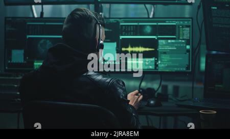 Cheerful male hacker in headset rejoicing in a successful cyber attack in a dim room of cybercriminals with computer equipment and software Stock Photo