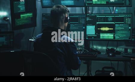 Cheerful male hacker in headset rejoicing in a successful cyber attack in a dim room of cybercriminals with computer equipment and software Stock Photo