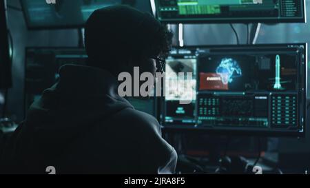 Young male hacker in hat and glasses using computers to hack nuclear warhead and start war while sitting at table in dark room Stock Photo
