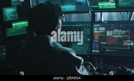 Young hacker drinking hot beverage and checking malicious code on computer monitors while sitting at desk in dark room at night Stock Photo