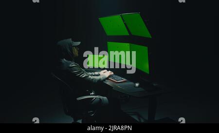 A male cybercriminal in glasses and a hood hacking the security system on the network and infecting with a virus in data on a computer with software, in a dark room at night Stock Photo